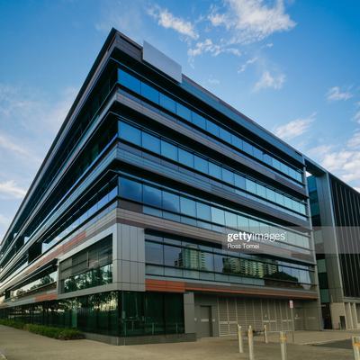 The image features a modern multi-story building with a sleek, angular design, characterized by extensive glass facades and a mix of dark and light materials. The structure is set against a clear blue sky, showcasing a contemporary architectural style.