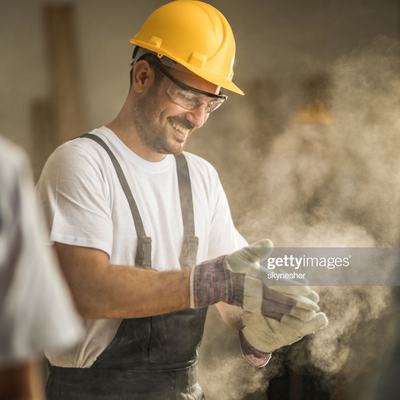 A smiling man in a yellow hard hat wears protective glasses and gloves, preparing for work in a dusty environment. He appears to be in a construction setting, focusing on his task as particles float around him. Other workers are blurred in the background.